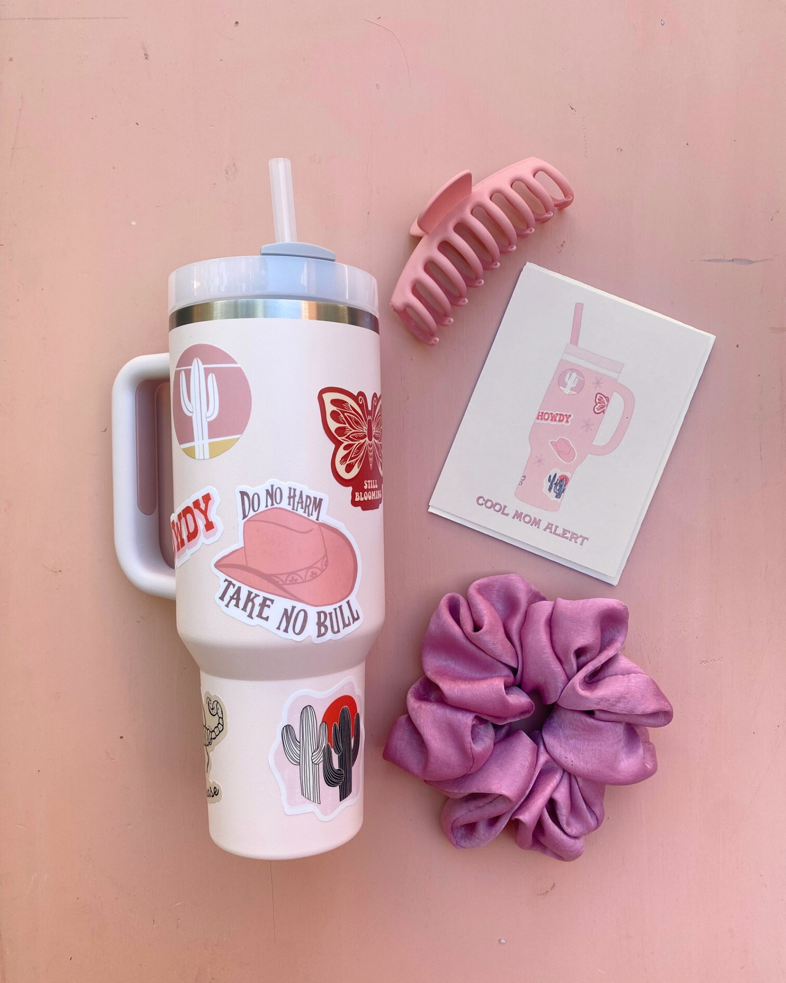 Flatlay photo of greeting card, stanley cup with stickers that is similar to illustrations on card, scrunchie, and hair clip