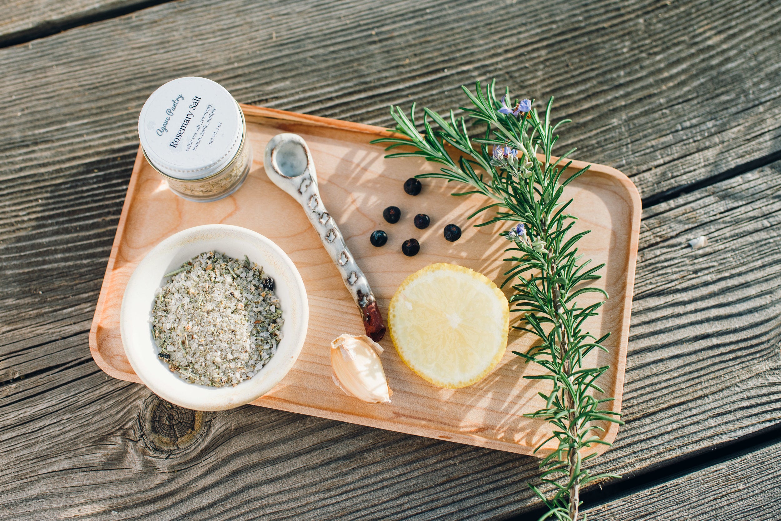 Light wood tray with rosemary salt in a jar and in a bowl next to a spice of lemon and a sprig of rosemary and a ceramic spoon