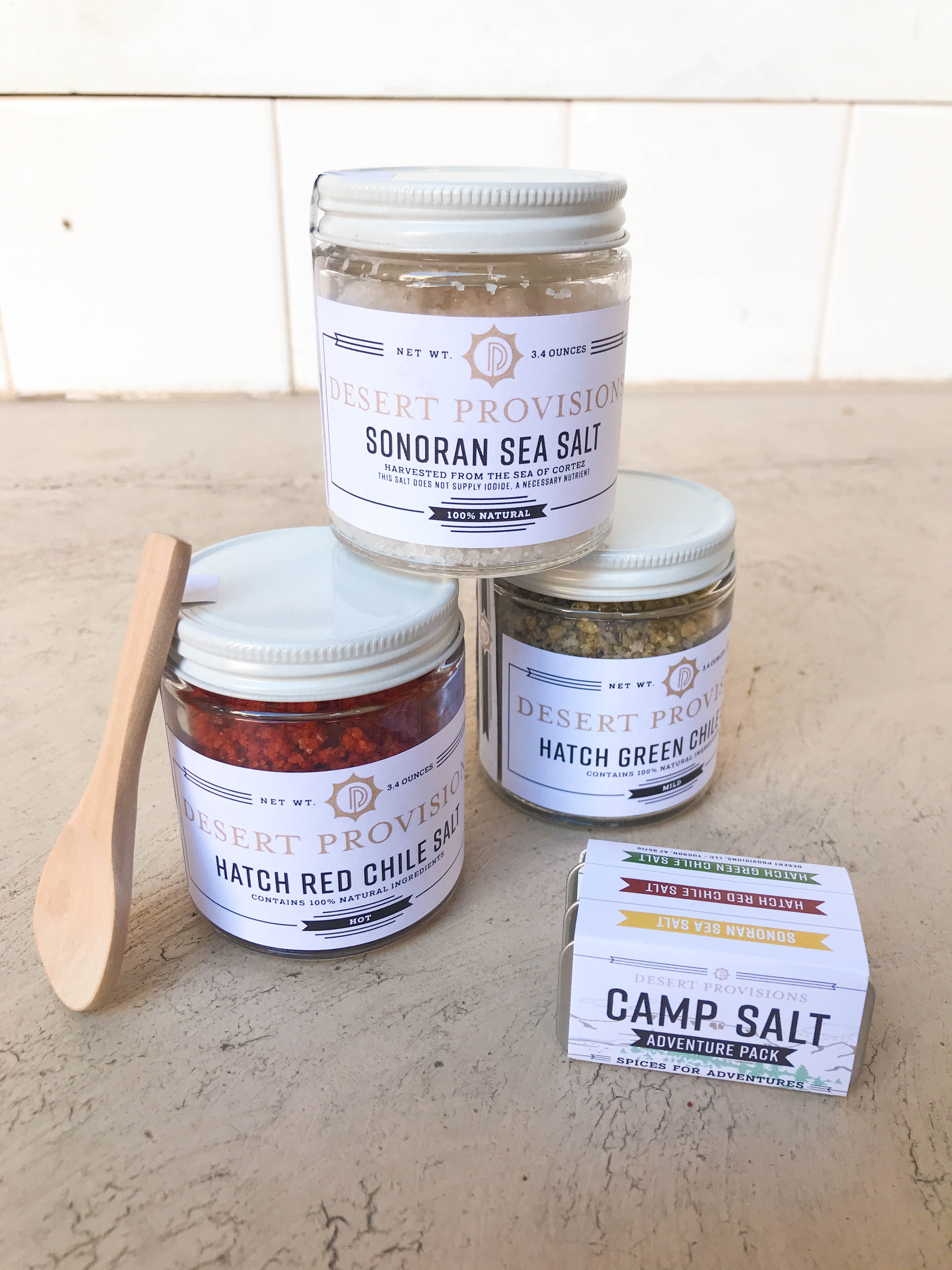 Stack of full size sonoran sea salt, hatch red chile salt, and hatch green chile salt next to mini camp salt packaging