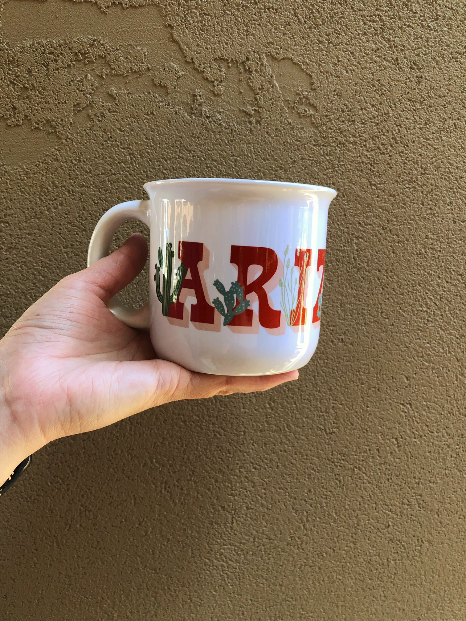 A hand holding an Arizona mug in front of a stucco wall