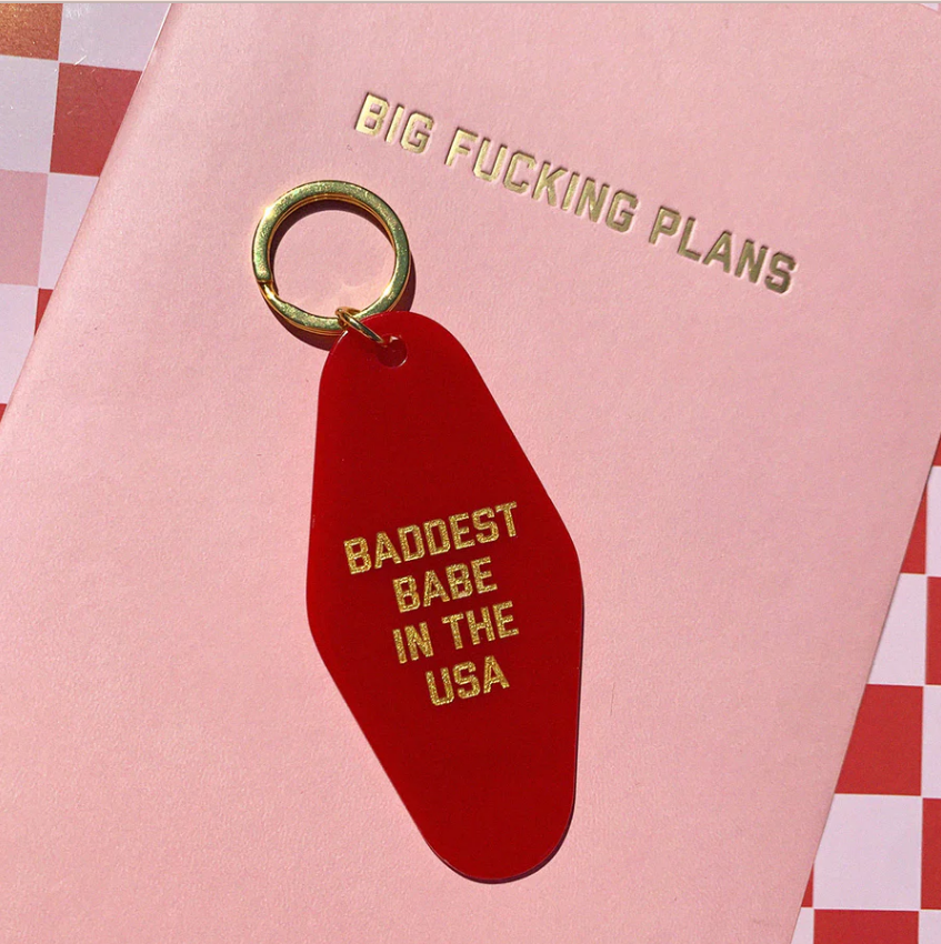 red motel style keychain reading baddest babe in the usa sitting on top of a journal that reads big fucking plans