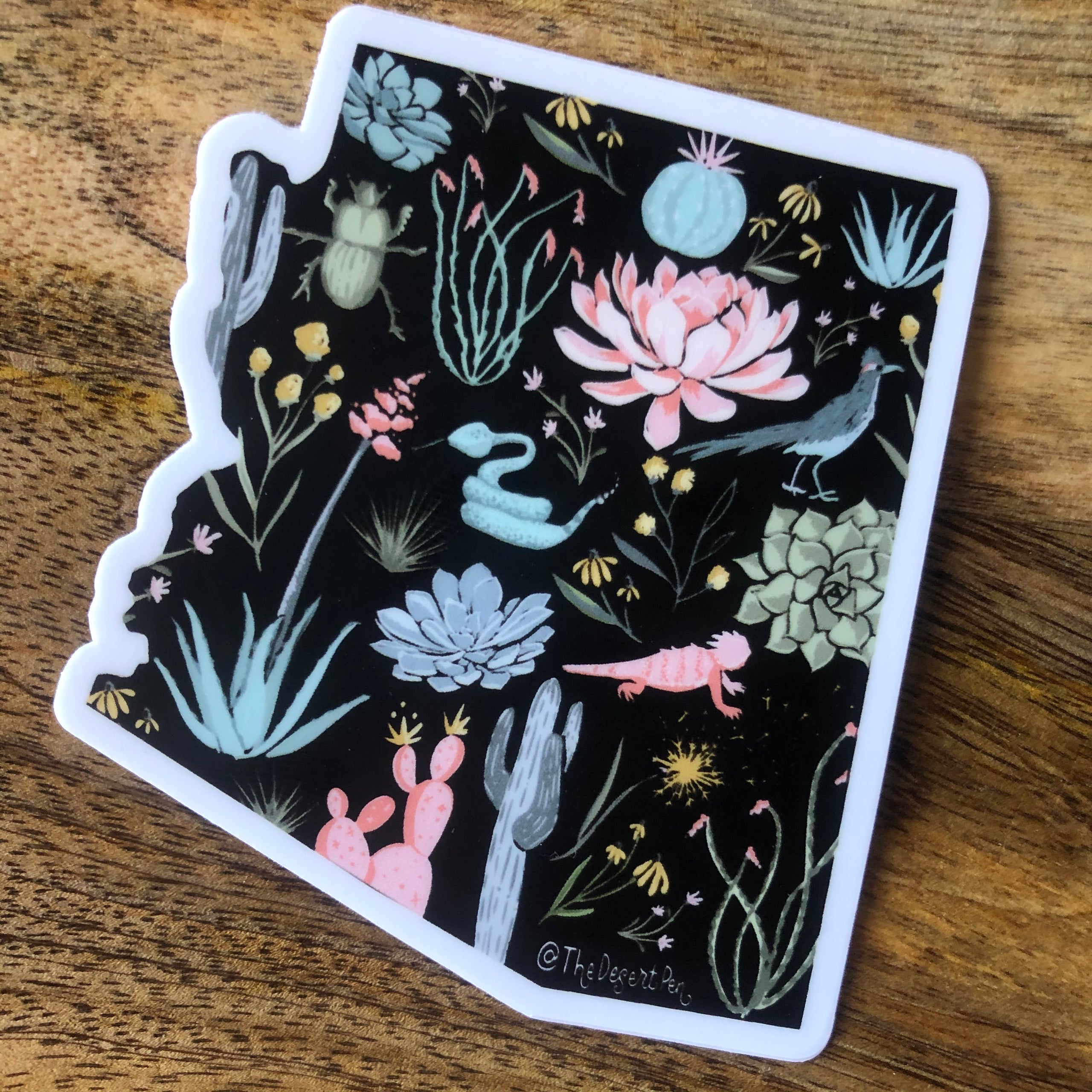 Black sticker shaped like arizona with illustrated plants and animals in pinks, greens, and grays with a wood background behind the sticker