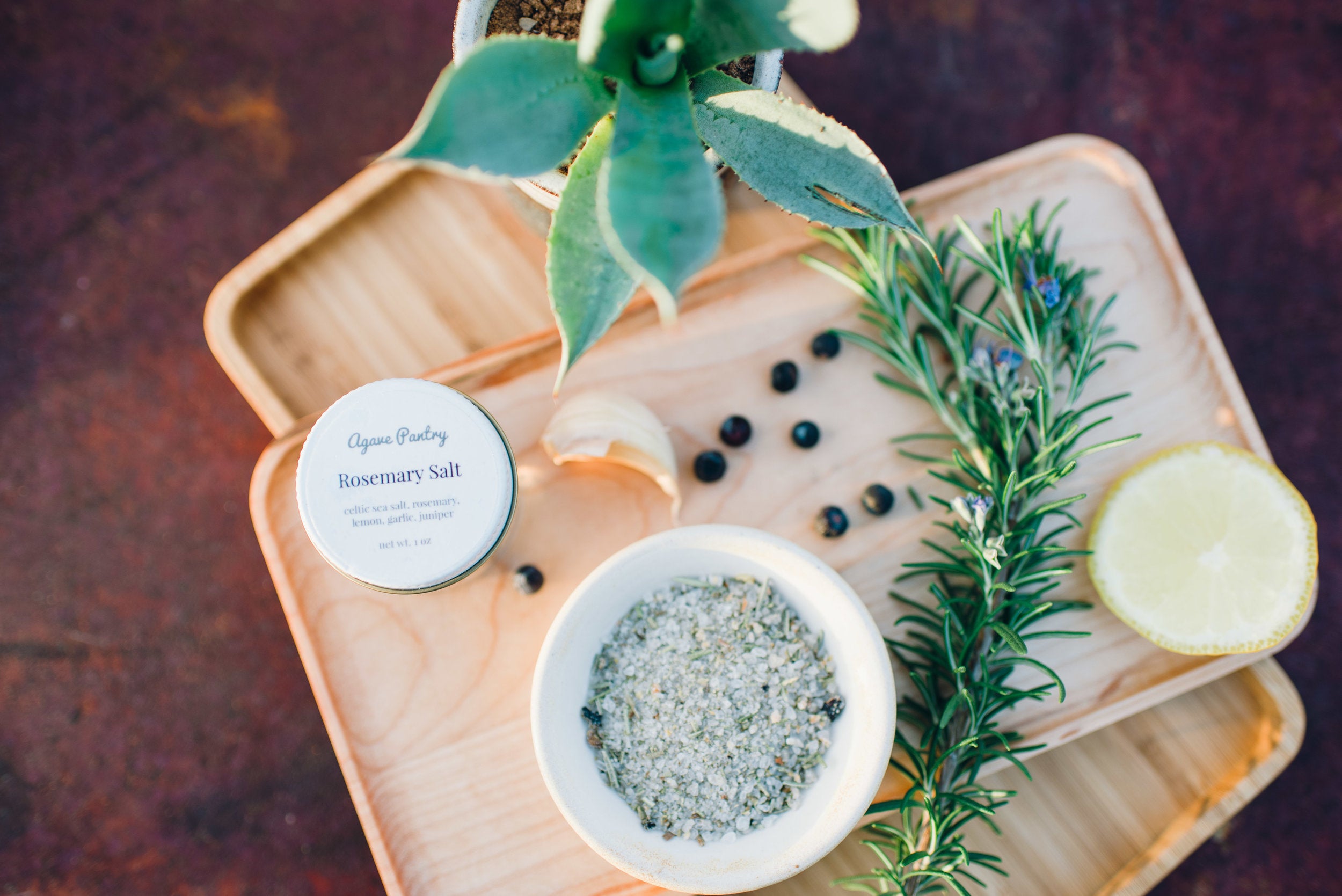 Light wood tray with rosemary salt in a jar and in a bowl next to a spice of lemon and a sprig of rosemary